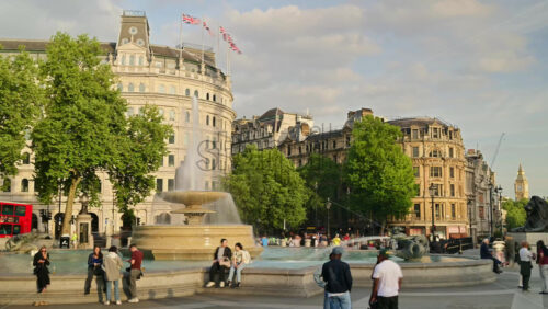 London, England – May 14, 2025: Time lapse of people enjoying a sunny afternoon in the Trafalgar Square with a water fountain, column, and moving red buses - Starpik Stock
