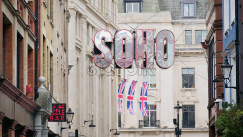 London, England – May 15, 2025: Close-up of the famous SOHO neon sign hanging over a street, with Union Jack flags fluttering in the background - Starpik Stock