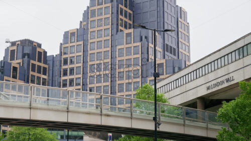 London, England – May 15, 2025: View of contemporary skyscrapers in the London Wall district with a sleek pedestrian bridge - Starpik Stock