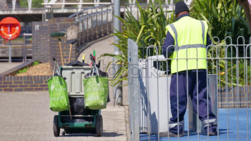 London, England – May 16, 2025: A street cleaner wearing a high-visibility vest, emptying a black garbage bag into a public litter bin - Starpik Stock