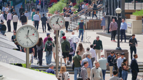 London, England – May 16, 2025: Busy pedestrian street filled with people walking, flanked by modern clocks on poles. Canary Wharf - Starpik Stock