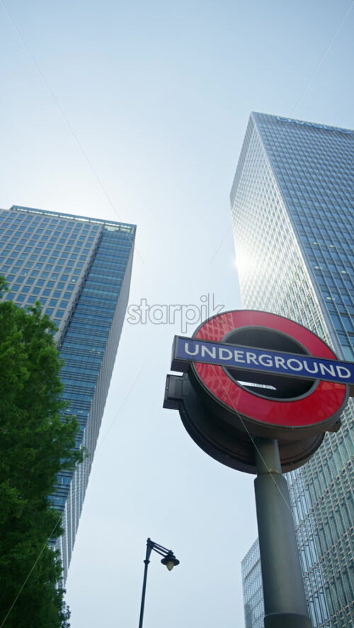 London, England – May 16, 2025: Iconic red Underground sign with skyscrapers on the background din daylight in Canary Wharf, London, England. Vertical - Starpik Stock