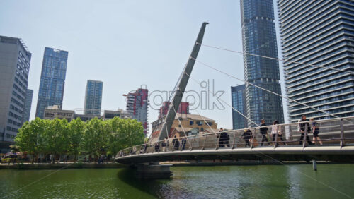 London, England – May 16, 2025: People walking across a modern pedestrian bridge over a canal in Canary Wharf, with residential and commercial high-rises in the background - Starpik Stock