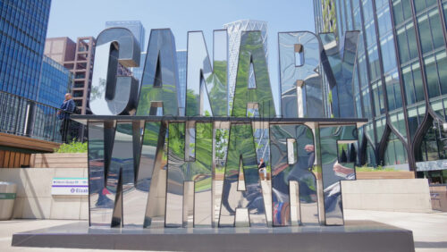 London, England – May 16, 2025: Reflective metallic sculpture spelling out Canary Wharf in a bustling plaza, surrounded by modern buildings and people walking or standing - Starpik Stock