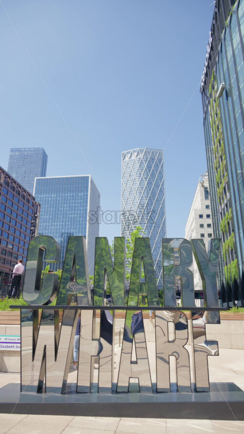 London, England – May 16, 2025: Reflective metallic sculpture spelling out Canary Wharf in a bustling plaza, surrounded by modern buildings and people walking or standing. Vertical - Starpik Stock