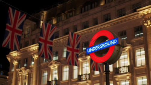 London Underground sign at night with illuminated British flags and classical building in the background in the evening - Starpik Stock