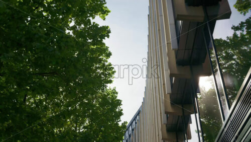 Low-angle shot of a modern office building framed by lush green trees and reflective window in London, England - Starpik Stock