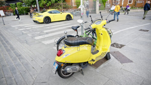 Milan, Italy – March 20, 2025: Bright yellow Vespa Sprint scooter parked on the street with a yellow car and people moving on the background - Starpik Stock