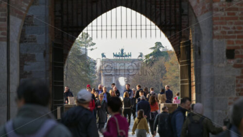 Milan, Italy – March 20, 2025: Multiple people entering and exiting the Sforzesco Castle in daylight with the Peace Arch in the background - Starpik Stock