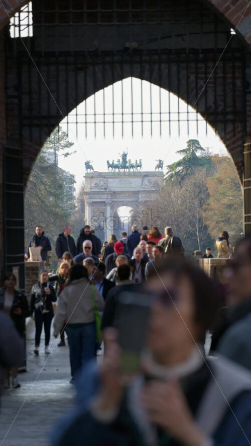Milan, Italy – March 20, 2025: Multiple people entering and exiting the Sforzesco Castle in daylight with the Peace Arch in the background. Vertical - Starpik Stock