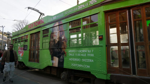 Milan, Italy – March 21, 2025: People walking by a green tram on the streets of the city in daylight - Starpik Stock