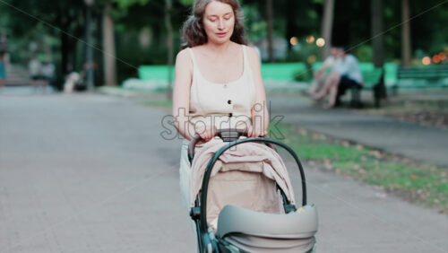 Mother dressed in light summer clothes pushing a baby stroller along a quiet park street - Starpik Stock