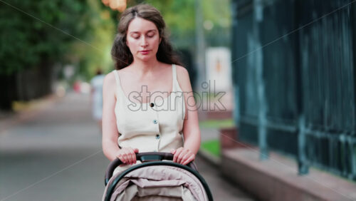 Mother dressed in light summer clothes pushing a baby stroller along a quiet park street - Starpik Stock