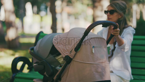 Mother sitting on a green bench in the park, gently rocking a stroller with her baby inside - Starpik Stock