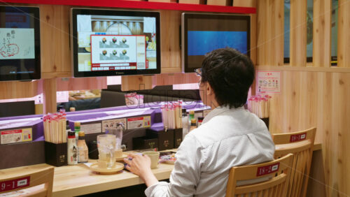 Nara, Japan – April 10, 2025: Man eating and ordering sushi from a digital menu while sitting alone at a wooden table - Starpik Stock