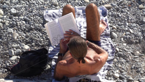 Nice, France – July 3, 2025: Overhead view of a man with long hair reading a book while sitting on a striped towel on the pebble beach - Starpik Stock