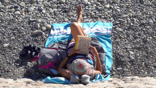 Nice, France – July 3, 2025: Overhead view of a woman lying on a beach towel on a pebbly beach, reading a codeword puzzle book under the summer sun - Starpik Stock
