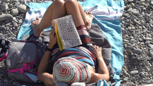 Nice, France – July 3, 2025: Overhead view of a woman lying on a beach towel on a pebbly beach, reading a codeword puzzle book under the summer sun - Starpik Stock