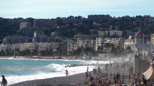 Nice, France – July 3, 2025: Waves crashing along the shore of the pebbled beach filled with people with the city’s skyline and palm-lined Promenade des Anglais in the background - Starpik Stock