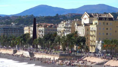 Nice, France – July 3, 2025: Wide view of the Promenade des Anglais with palm trees, historic buildings, and people enjoying the pebble shoreline - Starpik Stock