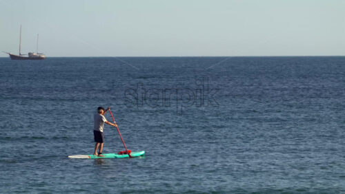 Nice, France – July 5, 2025: A man balancing on a stand-up paddleboard in calm coastal waters, with a large sailing ship visible on the horizon - Starpik Stock