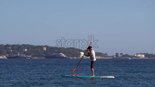 Nice, France – July 5, 2025: A man balancing on a stand-up paddleboard in calm coastal waters, with a large sailing ship visible on the horizon - Starpik Stock