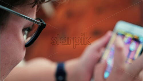Over-the-shoulder close-up of a kid with glasses playing on a smartphone indoors - Starpik Stock