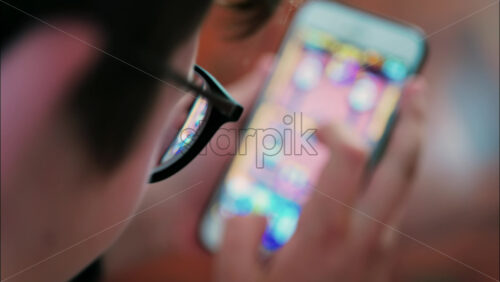Over-the-shoulder close-up of a kid with glasses playing on a smartphone indoors - Starpik Stock