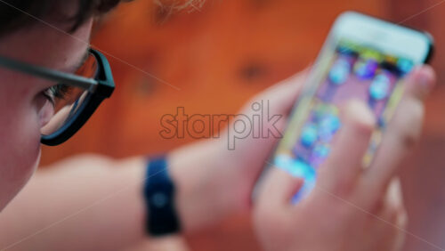 Over-the-shoulder close-up of a kid with glasses playing on a smartphone indoors - Starpik Stock