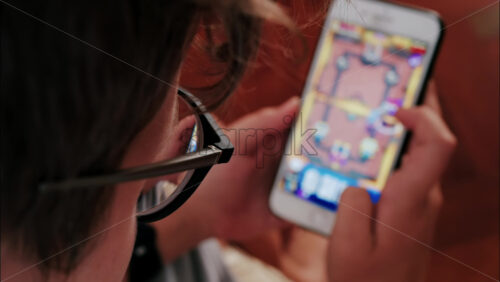 Over-the-shoulder close-up of a kid with glasses playing on a smartphone indoors - Starpik Stock