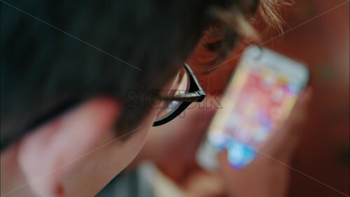 Over-the-shoulder close-up of a kid with glasses playing on a smartphone indoors - Starpik Stock