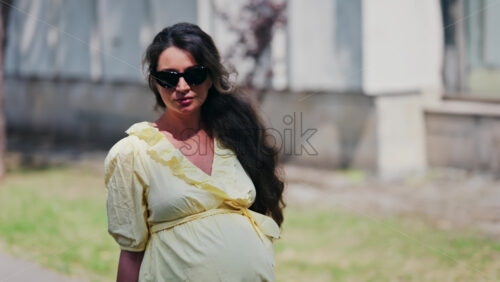 Pregnant woman in a yellow dress, wearing sunglasses walking on the street - Starpik Stock