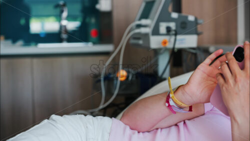 Pregnant woman scrolling on her phone while lying in a hospital bed at the doctor’s office - Starpik Stock