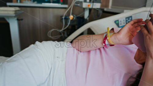 Pregnant woman scrolling on her phone while lying in a hospital bed at the doctor’s office - Starpik Stock