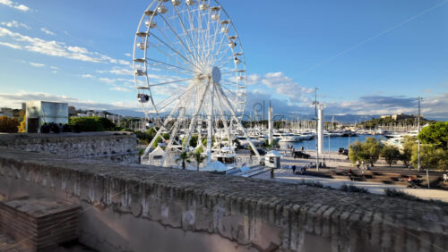 Side view of white ferris wheel rotating in Antibes, France with Port Vauban on the background - Starpik Stock
