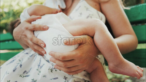 Tender moment of a mother breastfeeding her baby on a green park bench in daylight - Starpik Stock