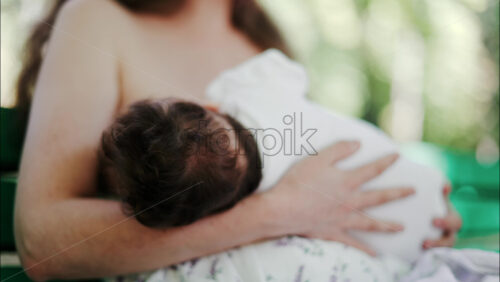 Tender moment of a mother breastfeeding her baby on a green park bench in daylight - Starpik Stock