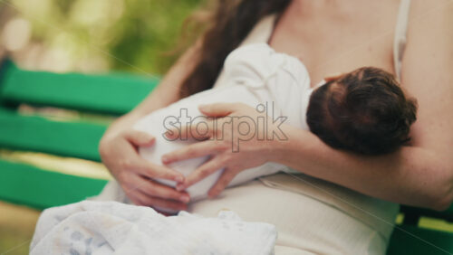 Tender moment of a mother breastfeeding her baby while sitting on a park bench - Starpik Stock