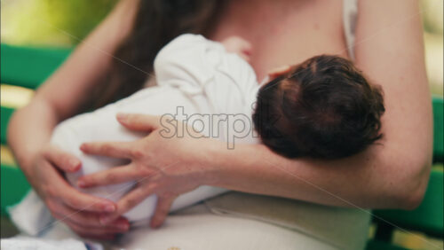 Tender moment of a mother breastfeeding her baby while sitting on a park bench - Starpik Stock