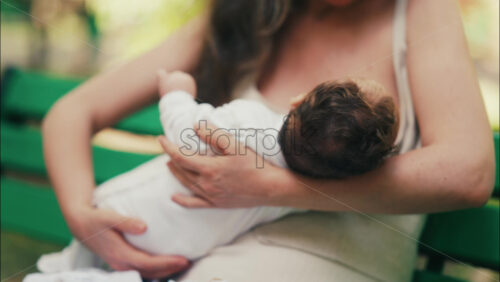 Tender moment of a mother breastfeeding her baby while sitting on a park bench - Starpik Stock