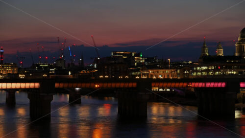 Time lapse of boats moving under the Thames with a lit bridge and the city skyline, including cranes and tall buildings i London, England at sunset - Starpik Stock