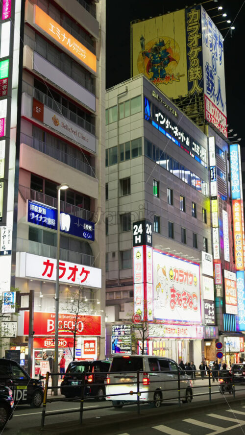 Tokyo, Japan – April 12, 2025: View of the vibrant life of Shinjuku City with people moving on the streets with lighted buildings in the evening. Translation: “Company names”. Vertical - Starpik Stock