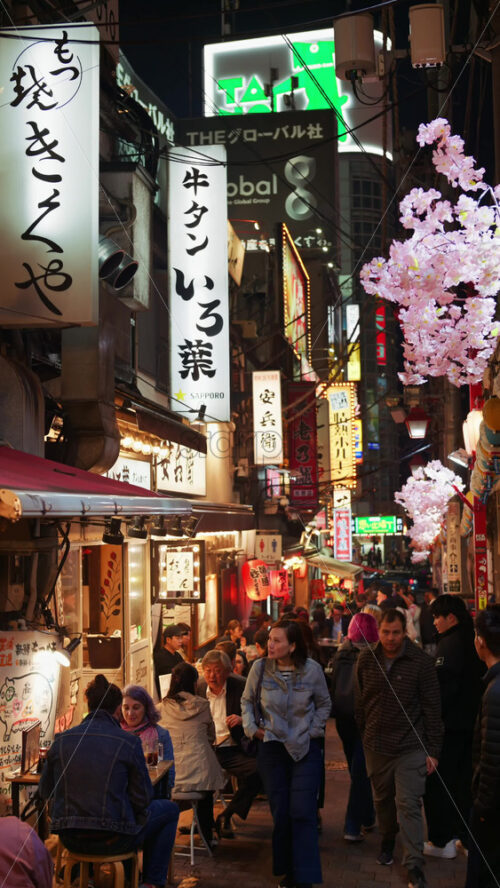 Tokyo, Japan – April 12, 2025: View of the vibrant life of Shinjuku City with people moving on the streets with lighted buildings in the evening. Translation: “Company names”. Vertical - Starpik Stock