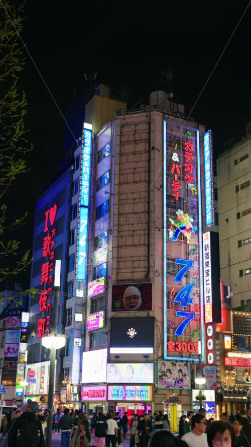 Tokyo, Japan – April 12, 2025: View of the vibrant life of Shinjuku City with people moving on the streets with lighted buildings in the evening. Translation: “Company names”. Vertical - Starpik Stock