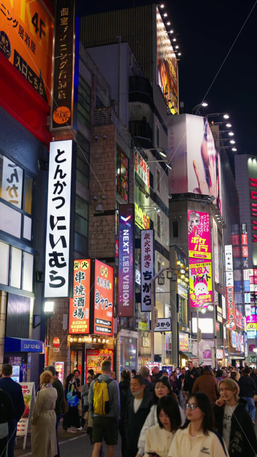 Tokyo, Japan – April 12, 2025: View of the vibrant life of Shinjuku City with people moving on the streets with lighted buildings in the evening. Translation: “Company names”. Vertical - Starpik Stock