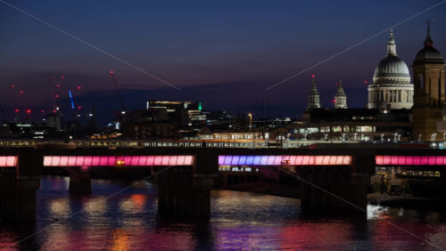 Trains moving on a bridge over the Thames with a lit bridge and the city skyline, including cranes and tall buildings i London, England at sunset - Starpik Stock