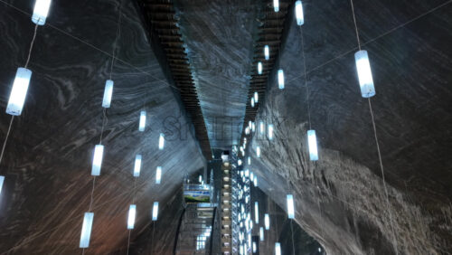 Turda, Romania – July 20, 2025: View of people walking through the underground amusement park inside the Turda salt mine, with rides, a Ferris wheel, and glowing blue lights suspended from the ceiling - Starpik Stock