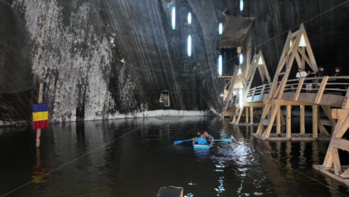 Turda, Romania – July 20, 2025: Visitors rowing small boats on a dark underground salt lake surrounded by wooden platforms and illuminated cavern walls at the Turda Salt Mine - Starpik Stock