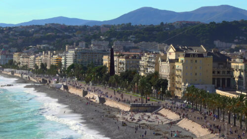 Turquoise waves crashing along the pebble beach of the Promenade des Anglais, with colorful buildings and palm trees lining the seafront - Starpik Stock