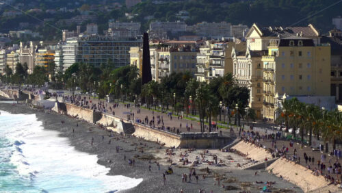 Turquoise waves crashing along the pebble beach of the Promenade des Anglais, with colorful buildings and palm trees lining the seafront - Starpik Stock